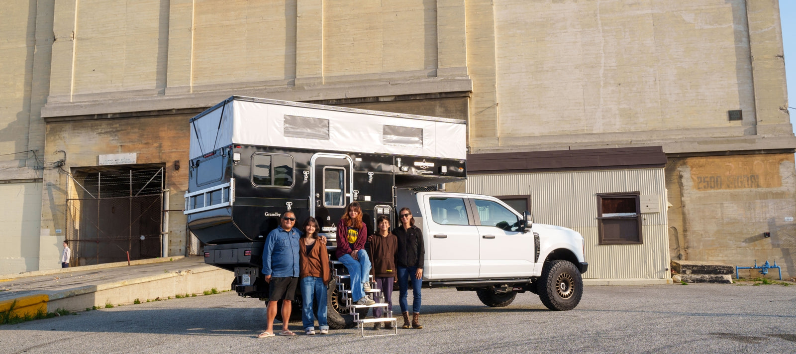 A white Ford F-350 with Bowen's custom flatbed truck and a pop-up Four Wheel Campers Grandby camper, with family together at the back of the truck in warm afternoon light.